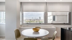 Ein moderner Essbereich mit einem runden Tisch, vier Stühlen, einer Schüssel und Blick auf eine Stadtkulisse durch große Fenster.