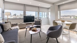 Ein modernes Wohnzimmer mit Flachbildfernseher, bequemen Sitzgelegenheiten, einer Essecke und großen Fenstern mit Blick auf die Stadt.