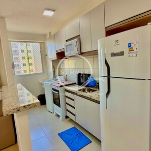 Une cuisine compacte avec des appareils électroménagers blancs, un four à micro-ondes et un réfrigérateur. Une fenêtre offre une vue sur les bâtiments environnants.