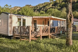 A row of mobile homes with wooden decks and large windows, set in a grassy area with trees and hills in the background. - Camping RCN La Bastide en Ardèche (Sampzon)