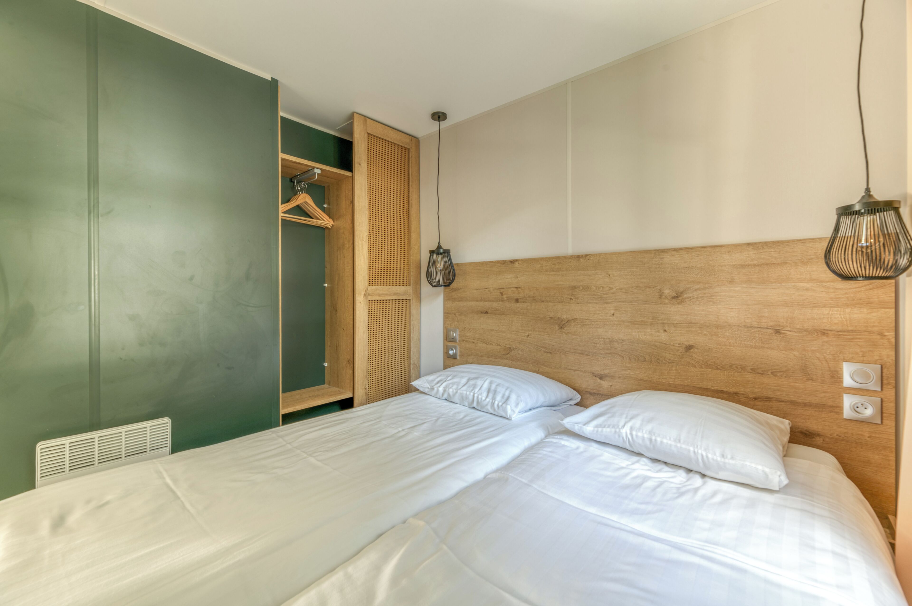 Een slaapkamer met een houten hoofdbord, twee bedden met witte lakens, een hanglamp en een groene muur met een inloopkast.