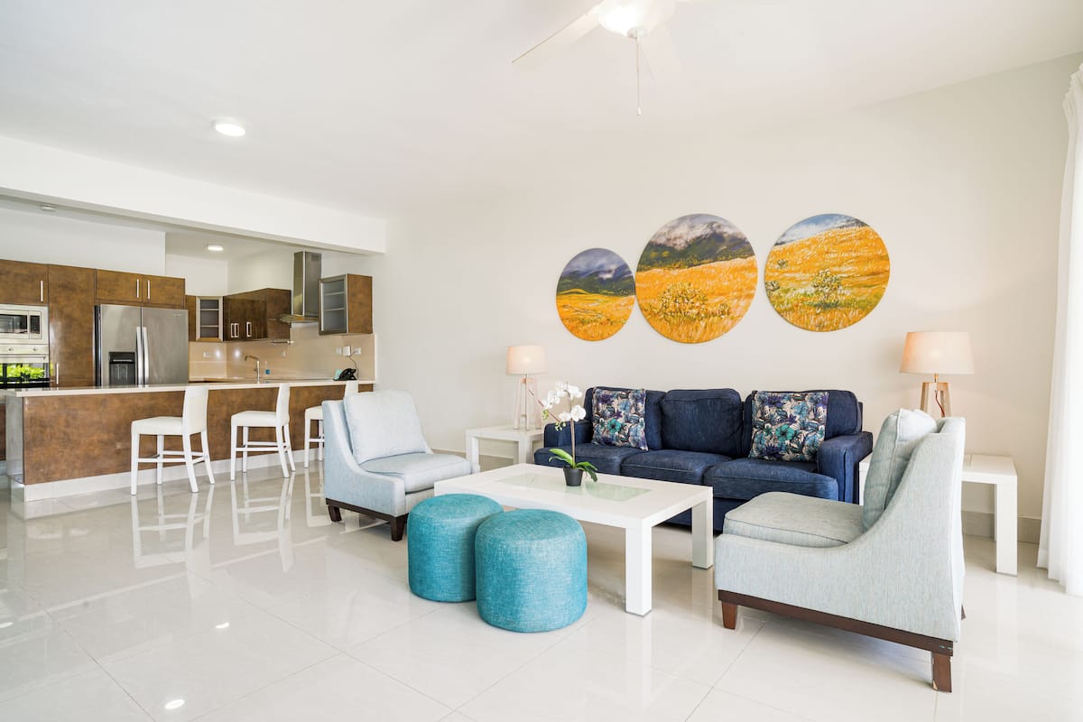 A modern living room with a sofa, ottomans, and a coffee table. There is a kitchen area with a refrigerator and cabinets.