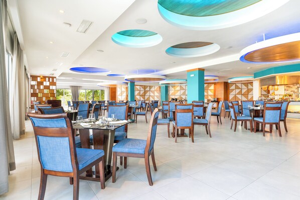 A spacious dining area with multiple tables and chairs, featuring a modern ceiling design with circular light fixtures. - Bakour Punta Cana Suites (Punta Cana)