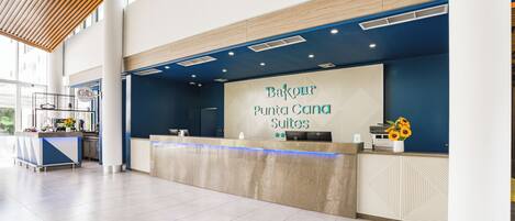 Un comptoir d’accueil avec une enseigne indiquant « Bakour Punta Cana Suites », situé dans le hall moderne d’un hôtel, doté d’un plafond haut et de grandes fenêtres.