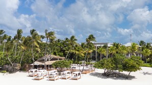 Un resort en la playa con sillas reclinables blancas, palmeras y un edificio con techo de paja.