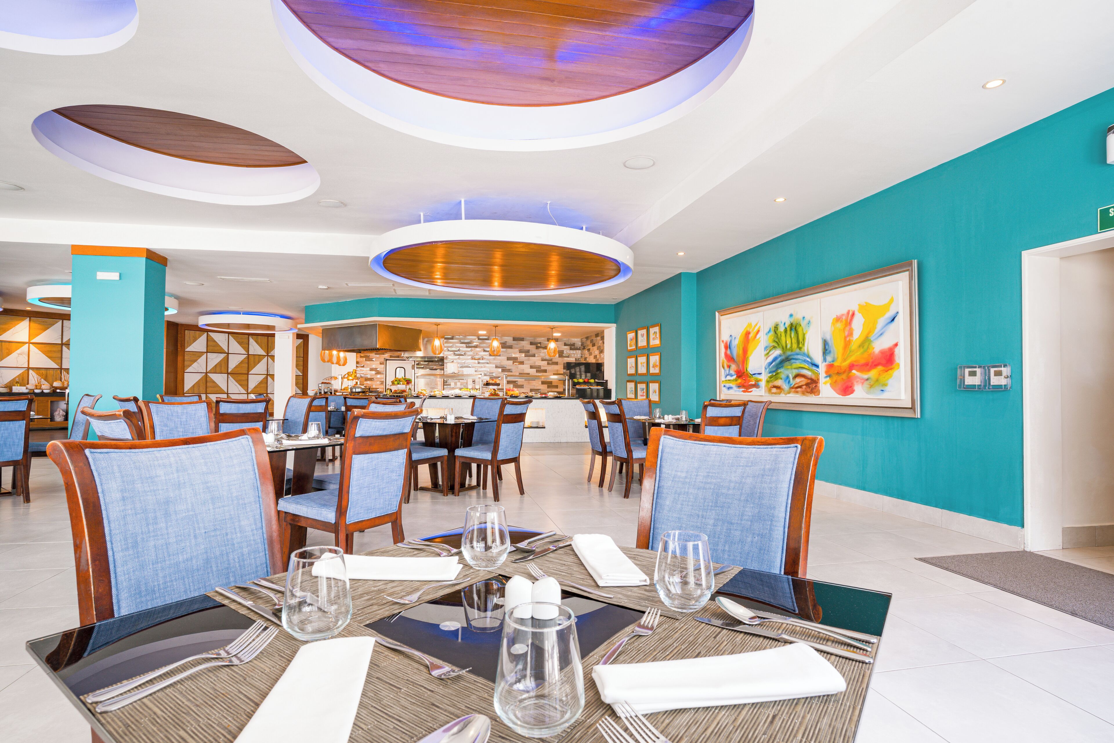 A modern restaurant with blue chairs, round tables, and a large painting on the wall.