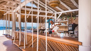 Un bar en la playa con terraza de madera, techo de paja y vista al océano.