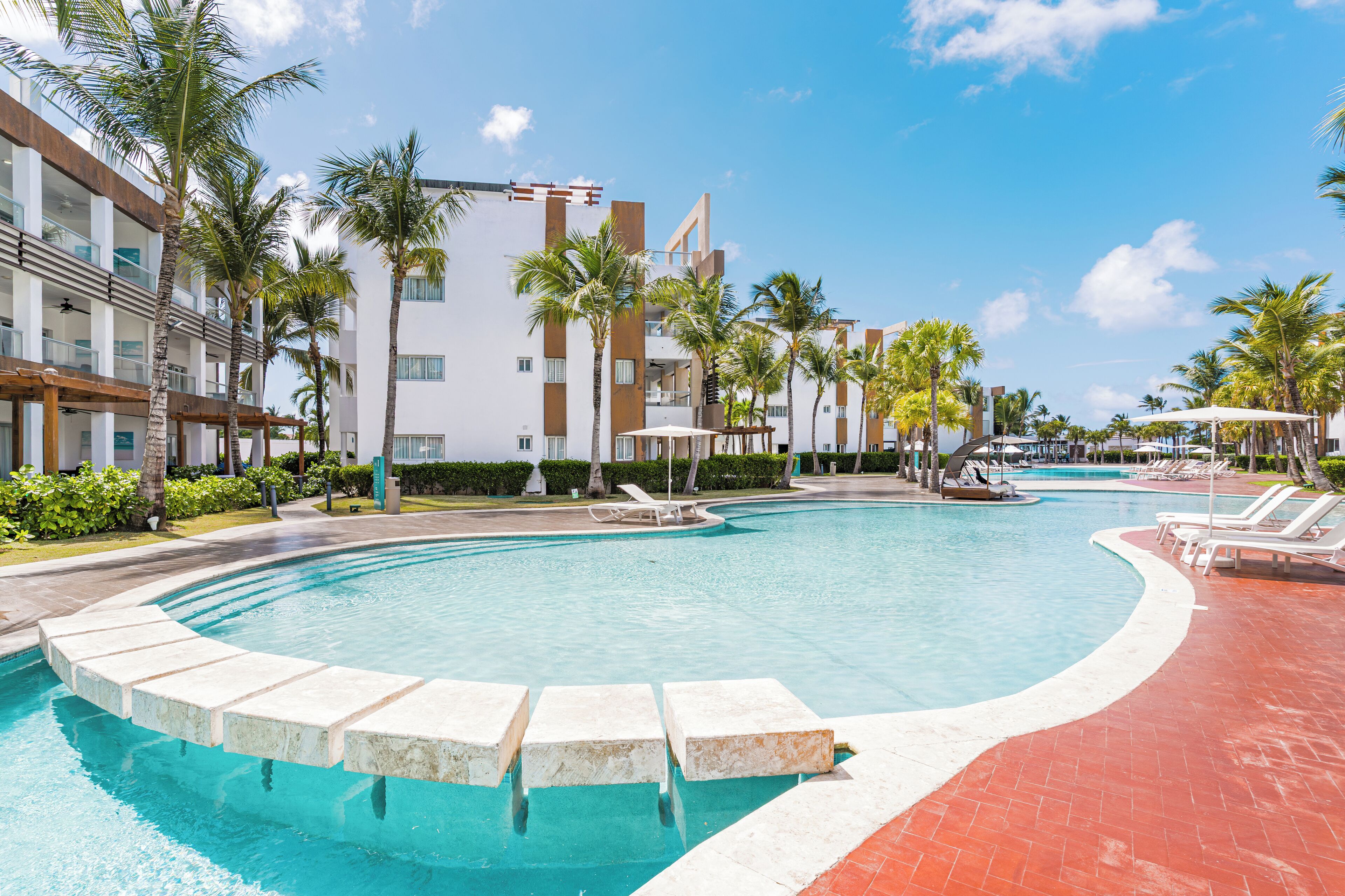 A swimming pool with lounge chairs, palm trees, and a modern building with balconies.