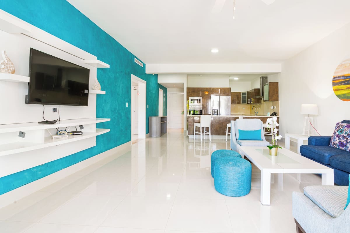 A modern living room with a flat-screen TV, a coffee table, and seating. A kitchen is visible in the background.
