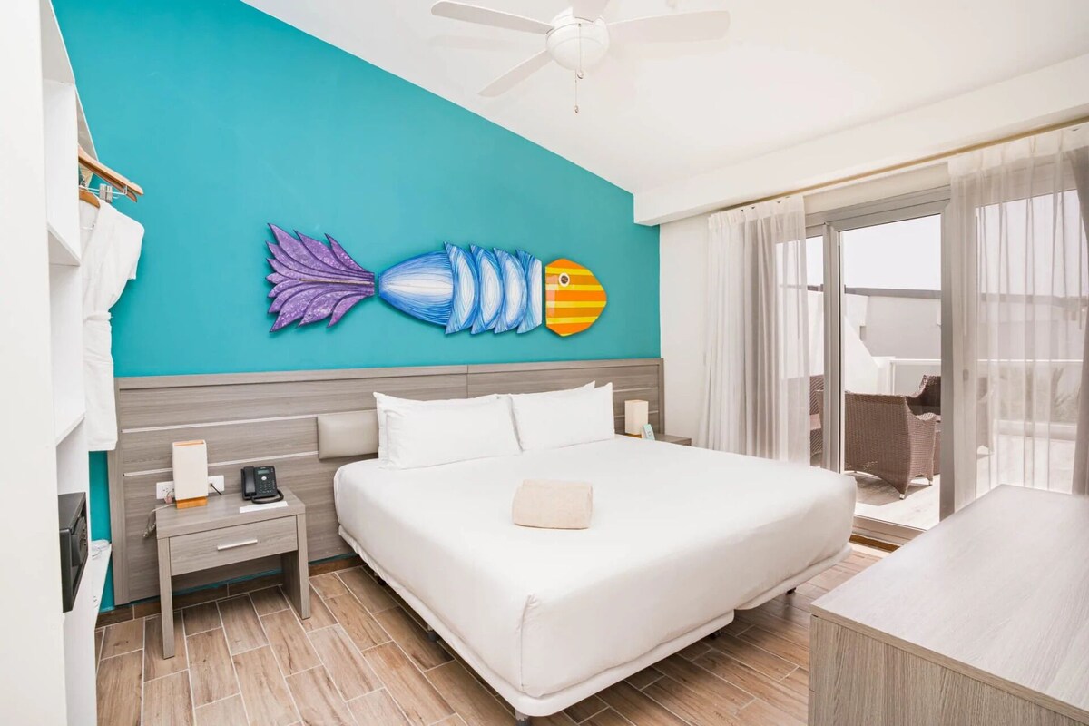 A hotel room with a bed, a nightstand, a wall art of fish, a ceiling fan, and a view of the outside.