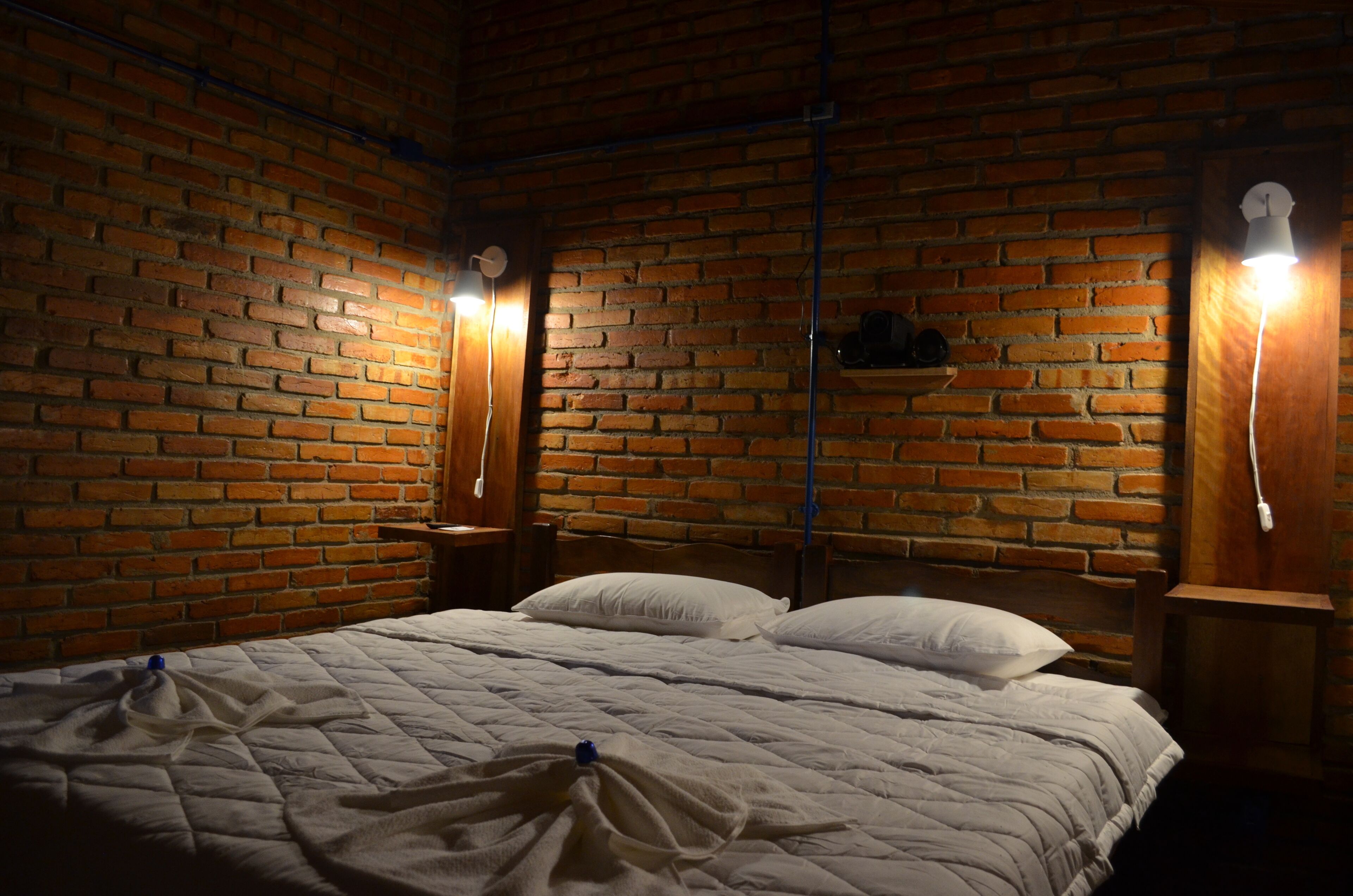 Quarto com parede de tijolos, cama com roupa de cama branca e duas luminárias de cabeceira.
