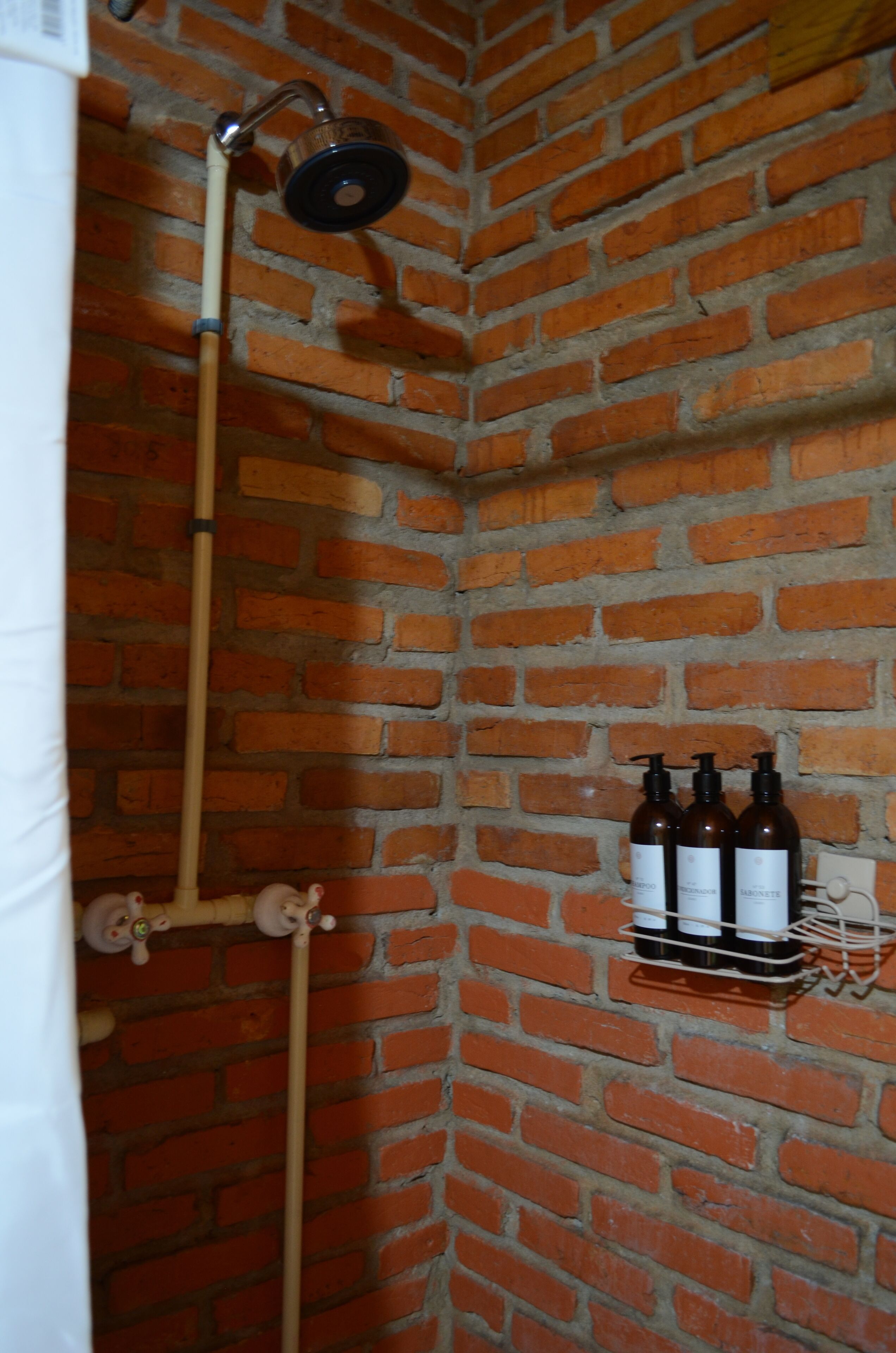A shower with a wooden handle and a brick wall. There are three bottles on a rack.