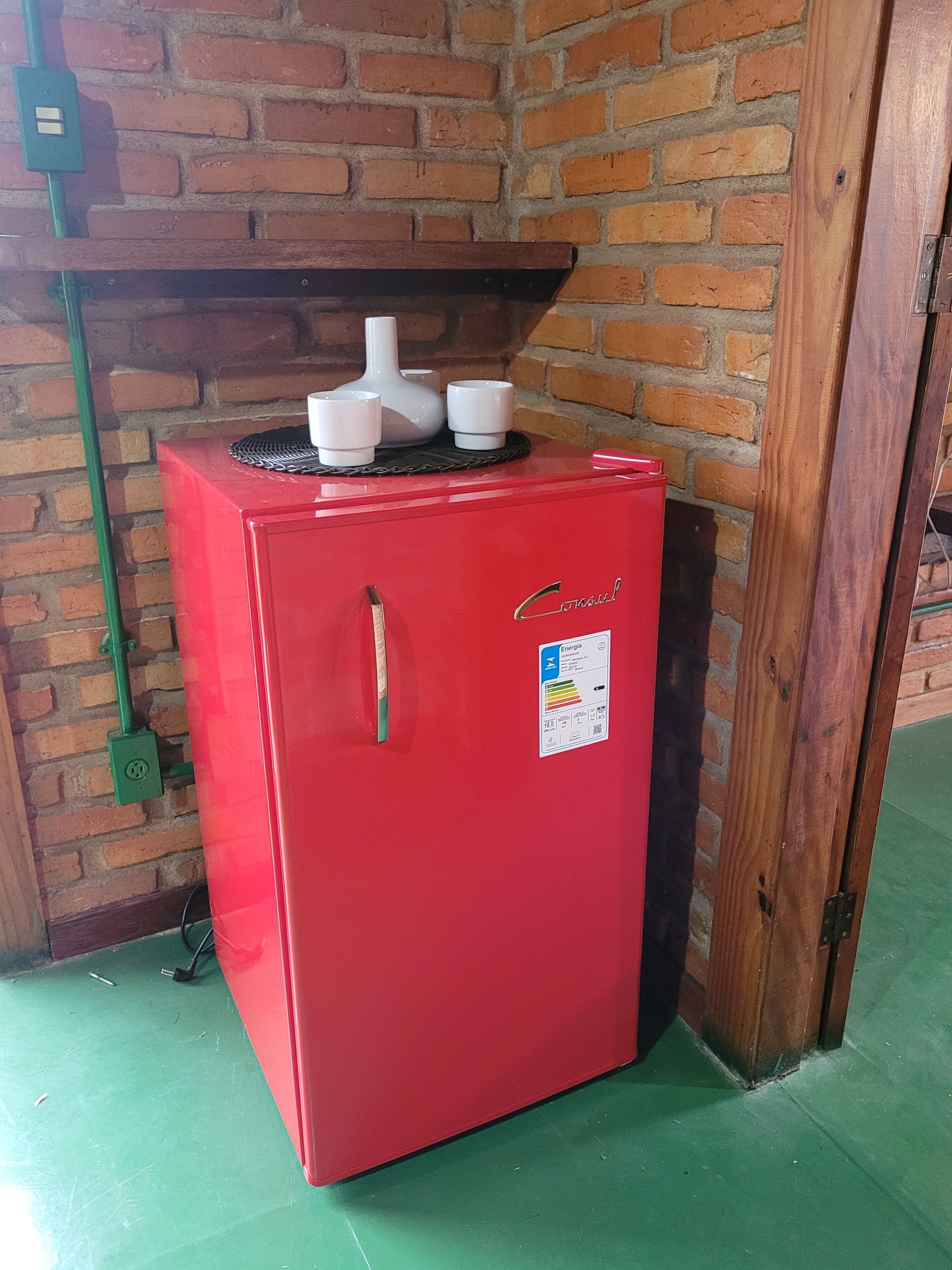 A red refrigerator with a white label, placed against a brick wall with a wooden door frame.