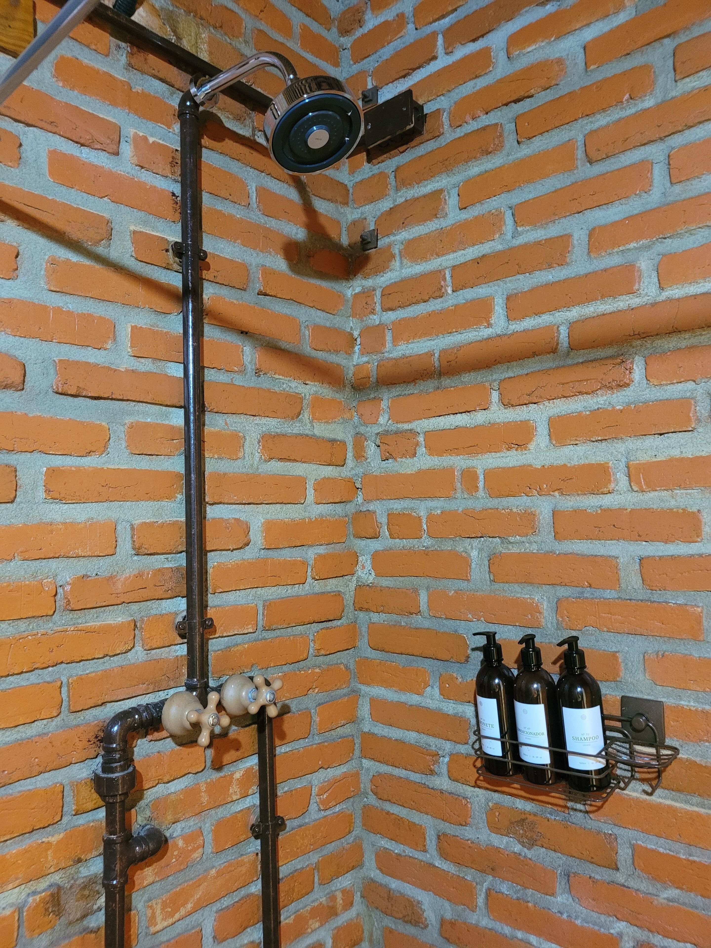 Esquina de una habitación con pared de ladrillo, una tubería negra y una estantería con botellas.