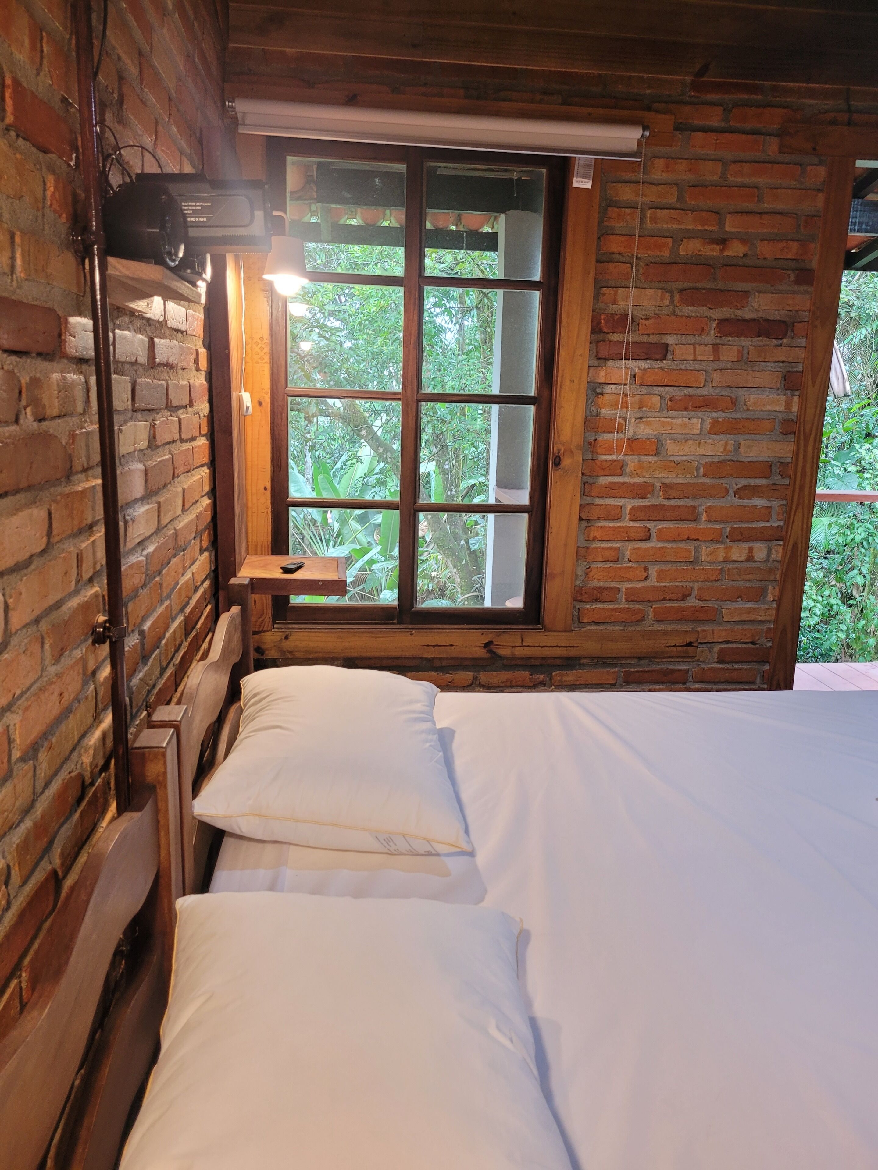 Quarto com estrutura de madeira, uma cama, janela com vista para vegetação e parede de tijolos.
