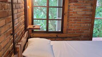 Quarto com estrutura de madeira, uma cama, janela com vista para vegetação e parede de tijolos.