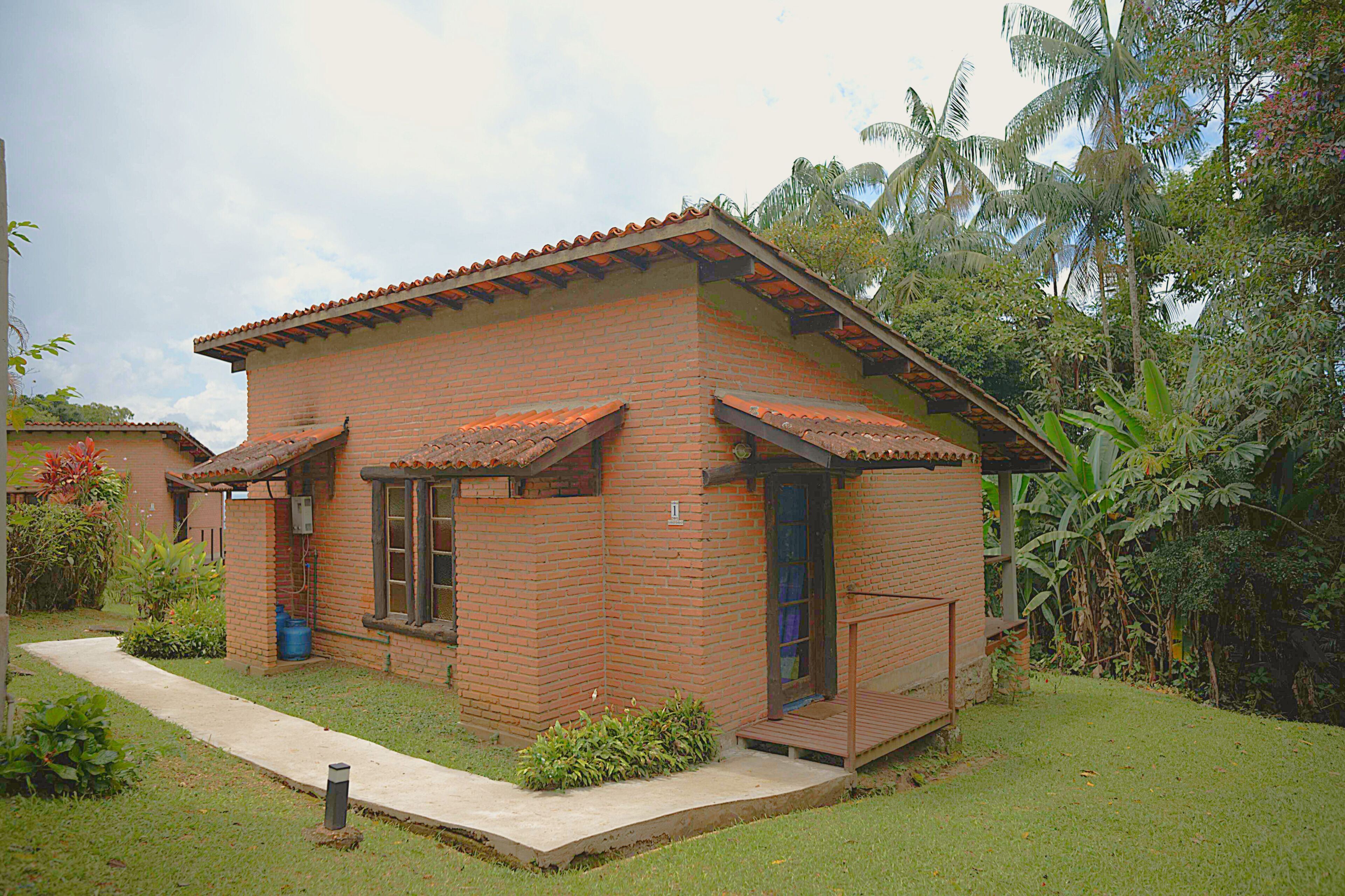 A small brick house with a tiled roof, surrounded by lush greenery and a clear sky.