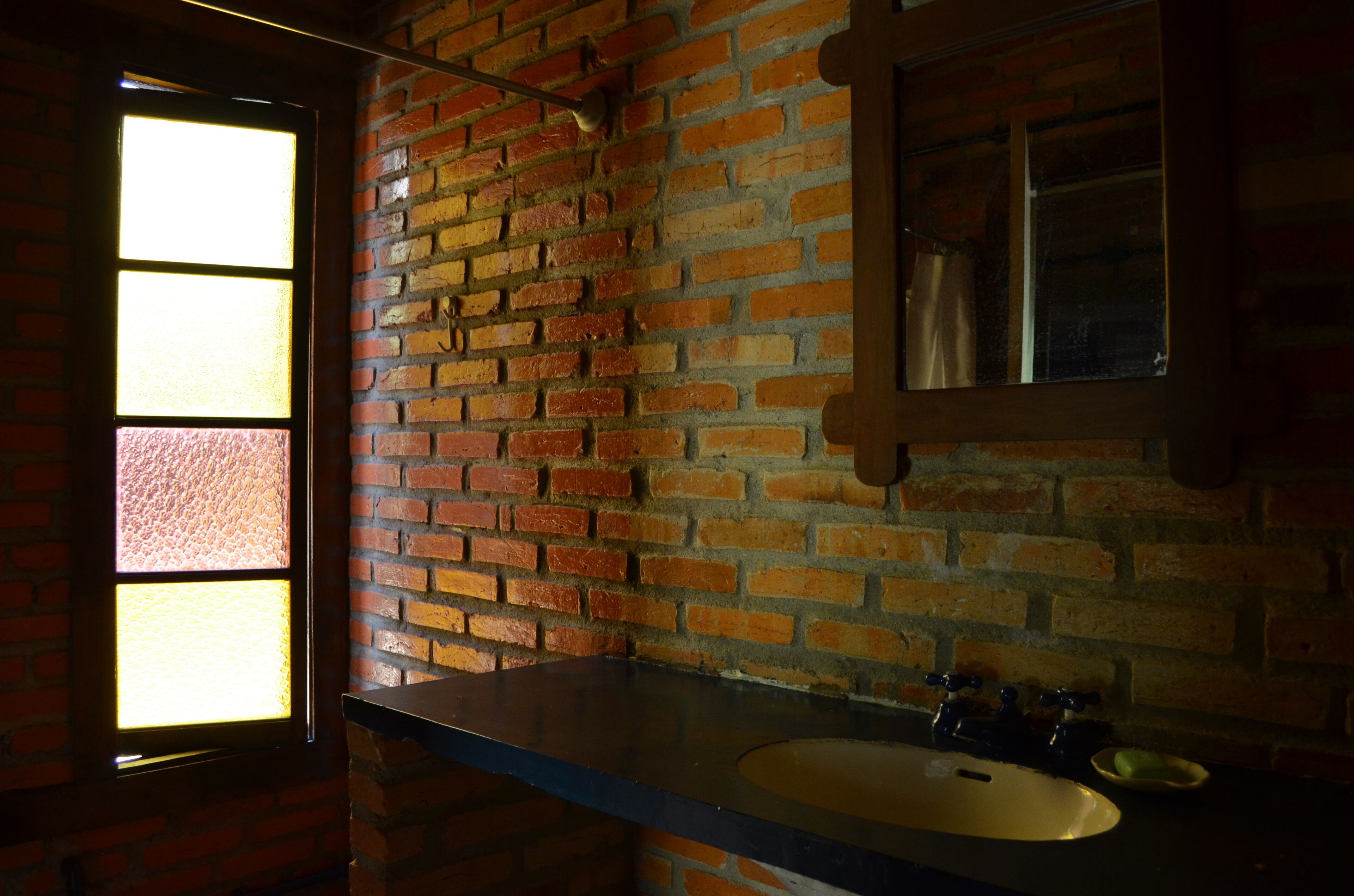 Un baño con pared de ladrillo, ventana, lavamanos y espejo.