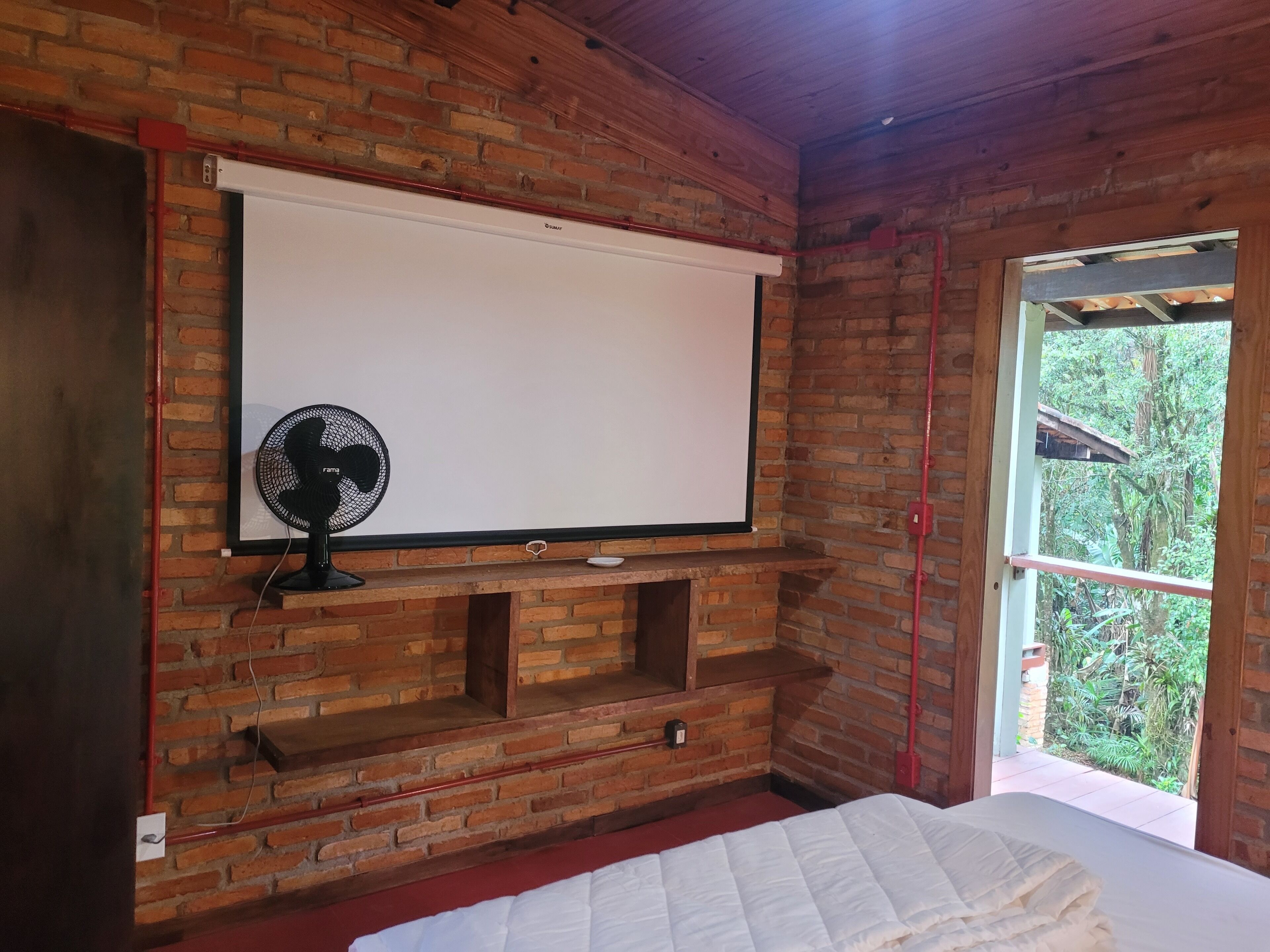 A room with a brick wall, a white bed, a small shelf, a fan, and a window with a view of greenery.
