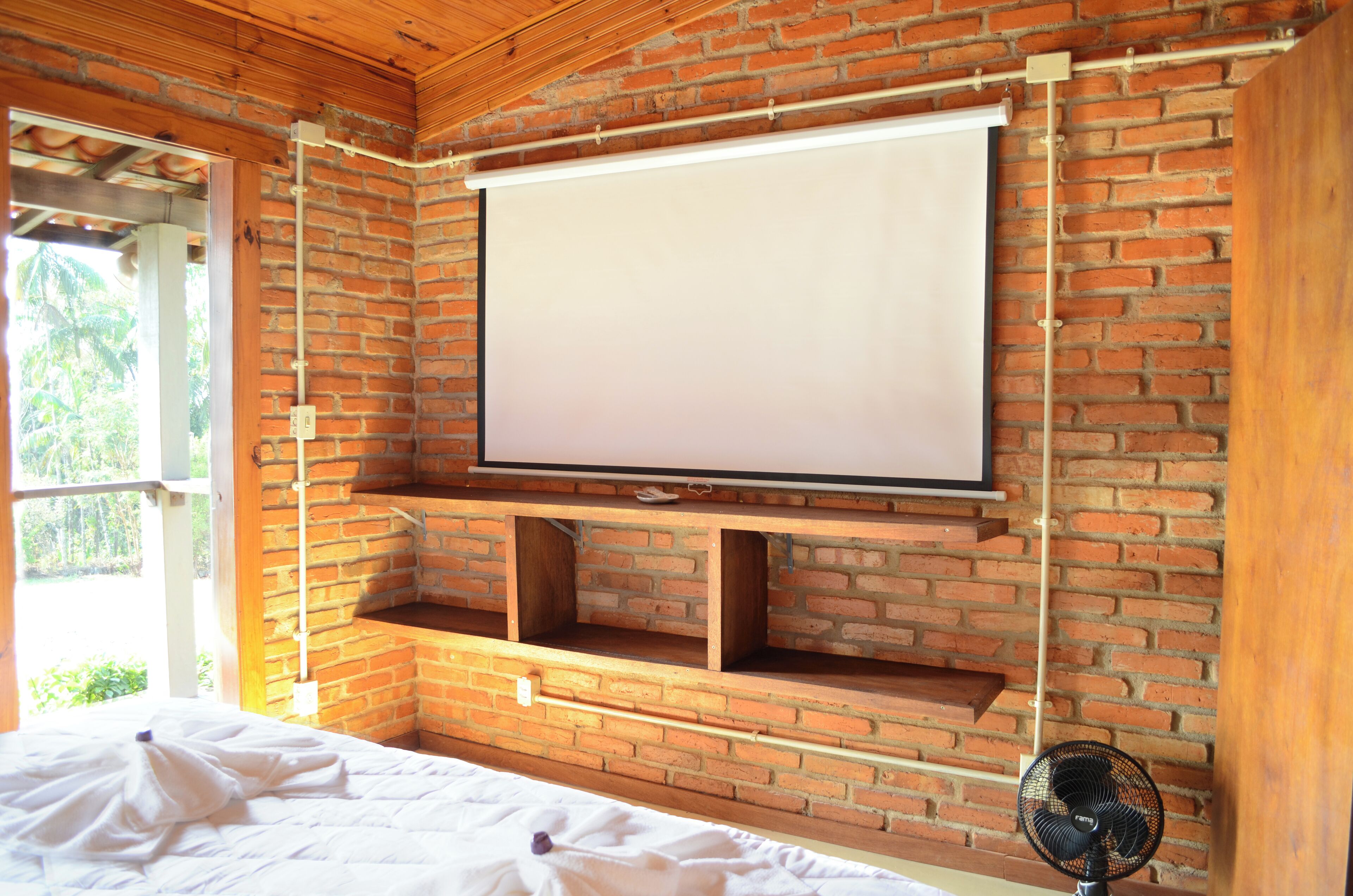 Quarto com parede de tijolos, tela de projeção fixada, estantes de madeira, cama e ventilador.