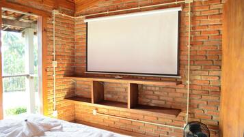 Quarto com parede de tijolos, tela de projeção fixada, estantes de madeira, cama e ventilador.