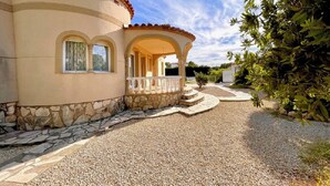 Una casa con base de piedra, fachada color beige y techo de tejas rojas. Un camino de grava conduce a un porche con barandilla blanca.