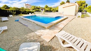 Una piscina rectangular con borde de piedra, rodeada por una zona de grava con sillas reclinables blancas.