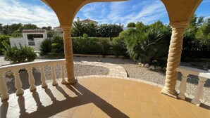 Un espacio al aire libre cubierto con suelo de baldosas, balaustradas blancas y vistas a un jardín con diversas plantas y un edificio al fondo.