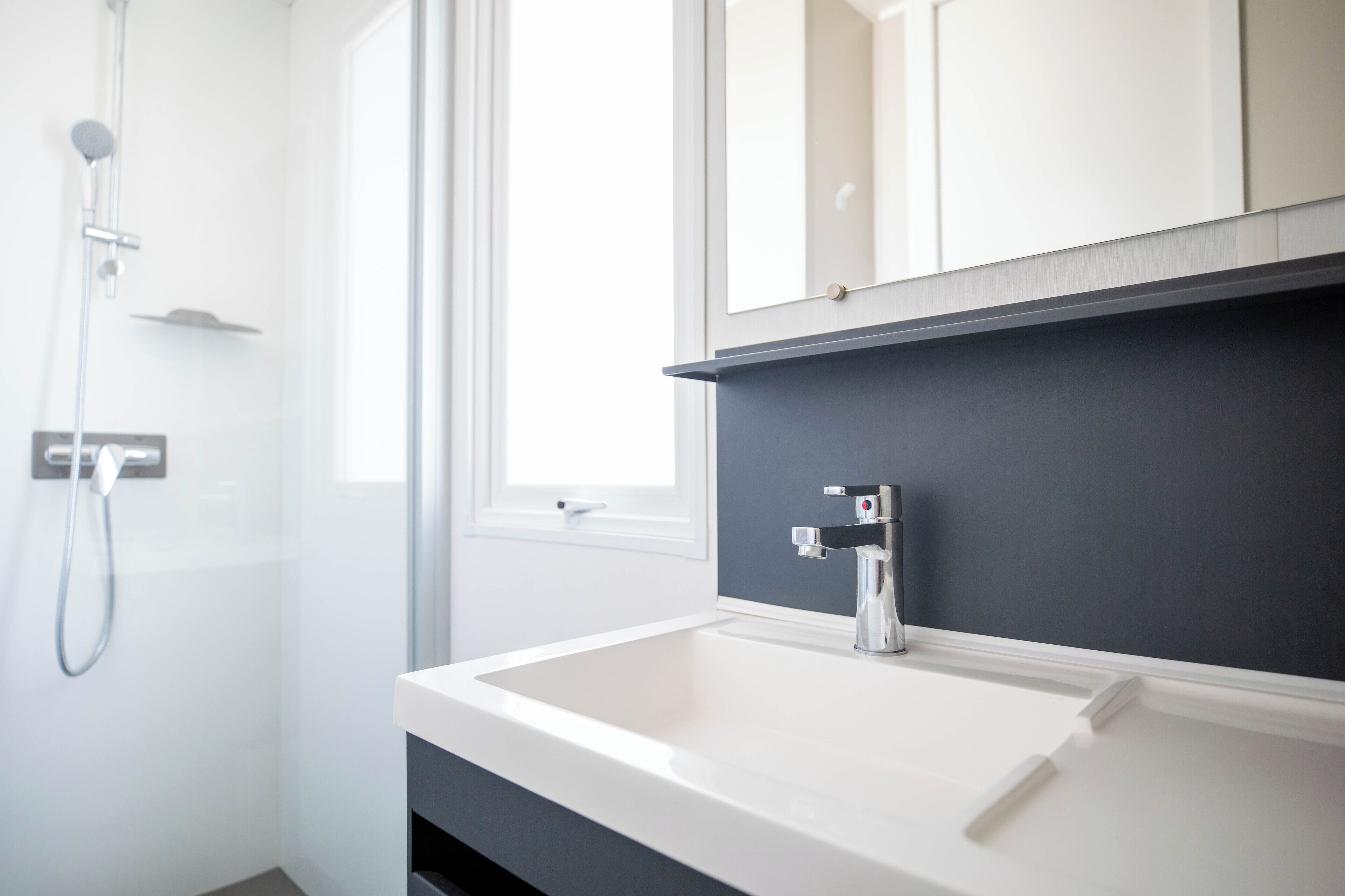 Une salle de bain moderne avec un lavabo blanc, un plan de toilette foncé et une douche équipée d’un pommeau de douche à main.