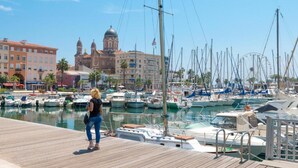 Un puerto deportivo con barcos amarrados, una mujer de pie en un muelle de madera y un edificio grande con cúpulas al fondo.