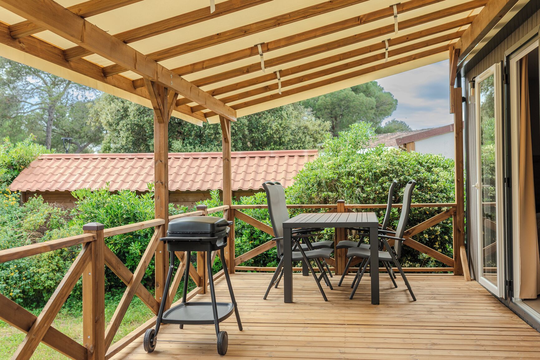 A covered outdoor deck with a table and chairs, a grill, and a view of a house with a red roof.