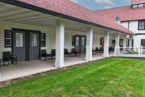 Une véranda couverte avec plusieurs portes et des chaises, une pelouse à l’avant et un bâtiment avec un toit rouge en arrière-plan.
