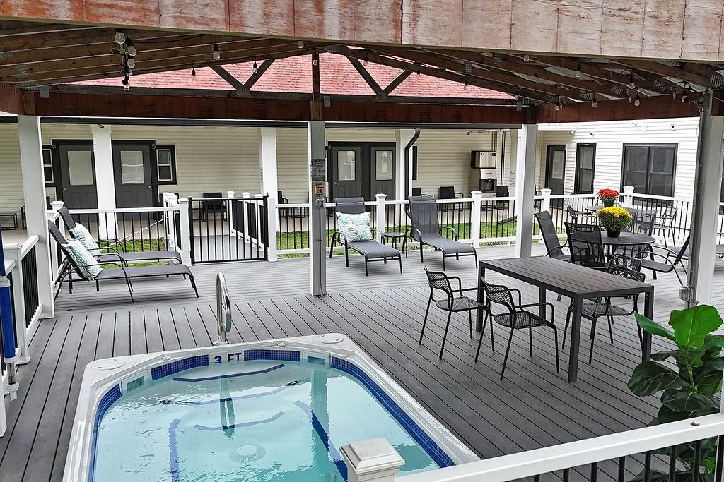 A covered outdoor deck with a hot tub, lounge chairs, and a table with chairs.
