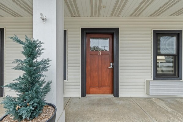 A house with a wooden front door, a small potted evergreen tree, and a window with blinds.