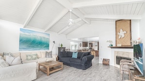 A spacious living room with a high ceiling, a large wave painting, a fireplace, and a kitchen in the background.