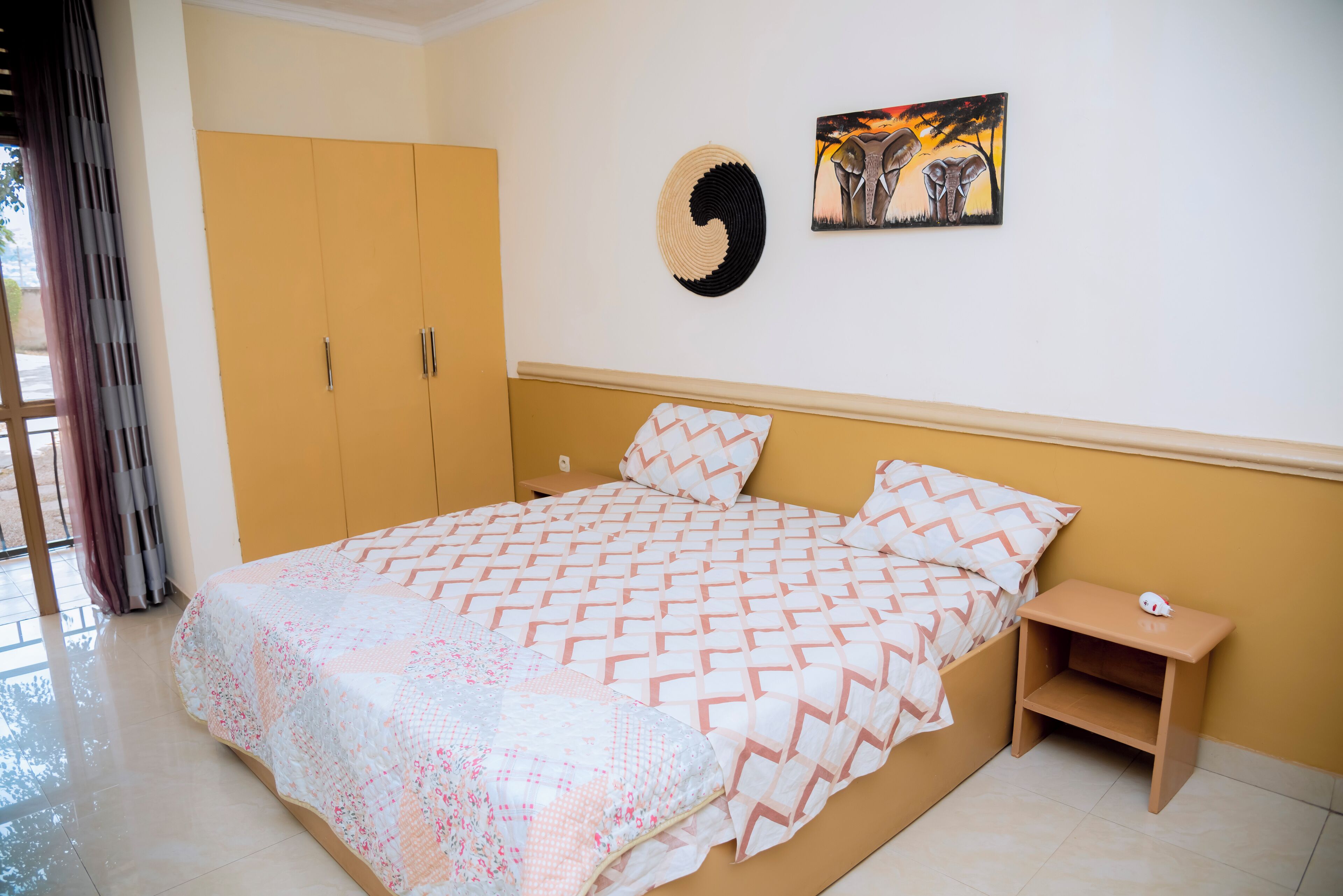 A bedroom with a bed, a wardrobe, a nightstand, a wall art piece, and a window with curtains.