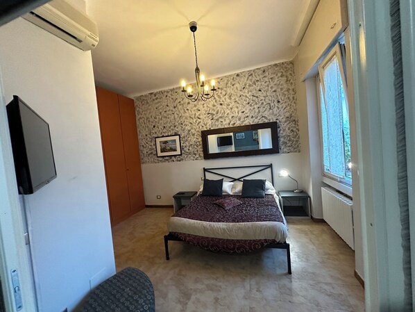 A bedroom with a bed, a chandelier, a TV, a mirror, and a window.