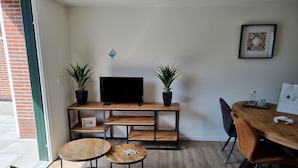 A modern living room with a TV on a wooden TV stand, two potted plants, a round coffee table, a dining table with chairs, and a framed picture on the wall. - 2B new vacation home with terrace and air conditioning (Westkapelle)