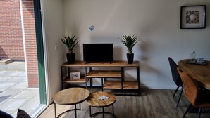 A modern living room with a TV on a wooden stand, two potted plants, a dining table with chairs, and a small framed picture on the wall. - 2B new vacation home with terrace and air conditioning (Westkapelle)