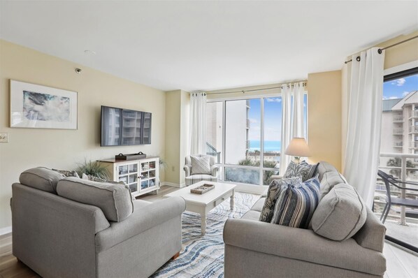 A living room with two sofas, a coffee table, a TV, and a cabinet. There is a large window with a view of buildings and a balcony.
