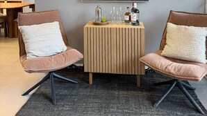 Un salon moderne avec deux fauteuils confortables, une table d’appoint en bois sur laquelle se trouvent une bouteille et des verres, et un grand tableau aux motifs tropicaux accroché au mur.