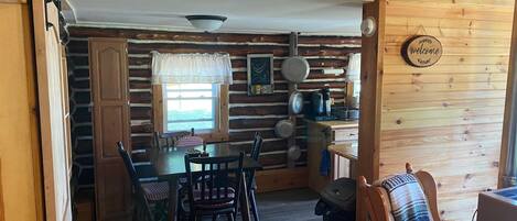 Interior rústico de una cabaña de madera con comedor, cocina con ollas colgantes y un letrero de bienvenida.