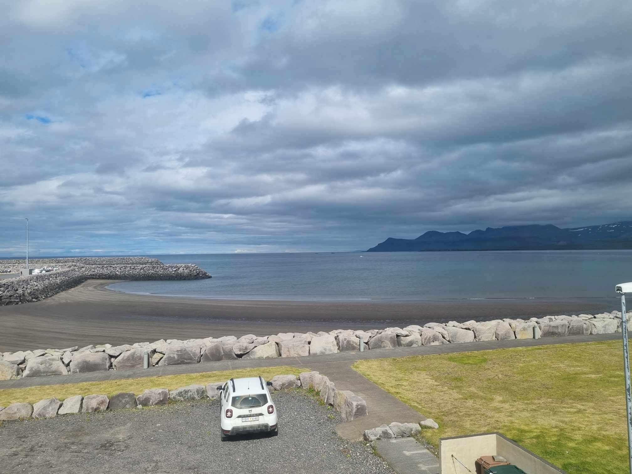Una carretera costera con un coche blanco estacionado al lado, un rompeolas rocoso y un mar tranquilo bajo un cielo nublado.