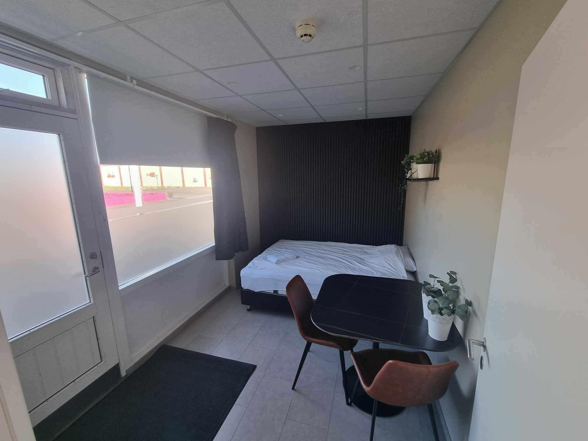 Une petite pièce, de taille modeste, comprenant un lit, une table et deux chaises. Une fenêtre offre une vue sur l’extérieur.