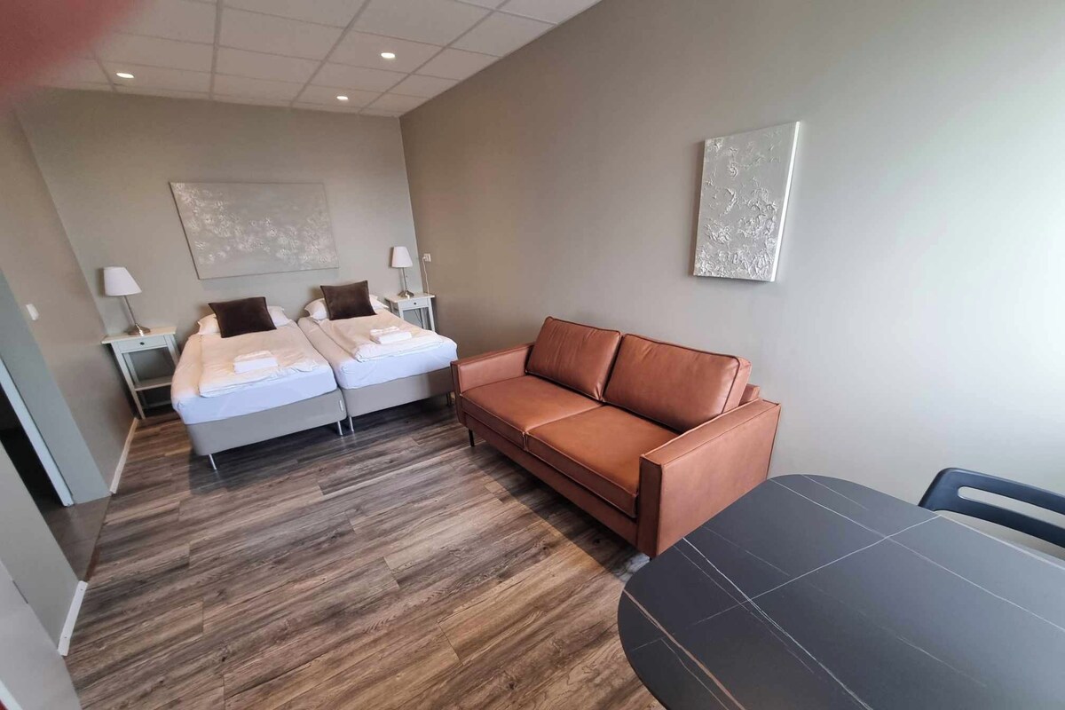 A modern hotel room with a wooden floor, a brown leather sofa, a bed with white linens, and a small table.