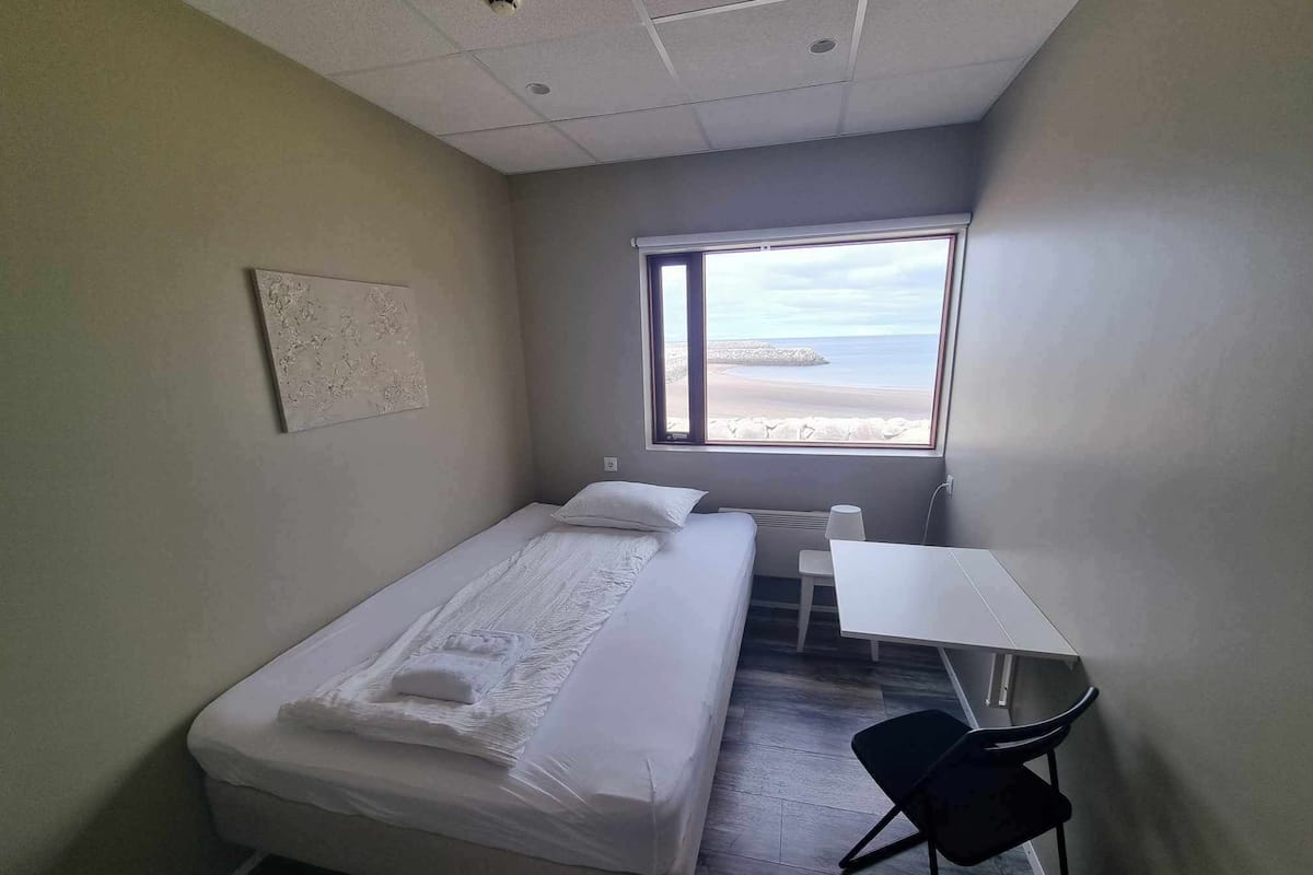 A small, clean room with a bed, a desk, and a window offering a view of the beach.