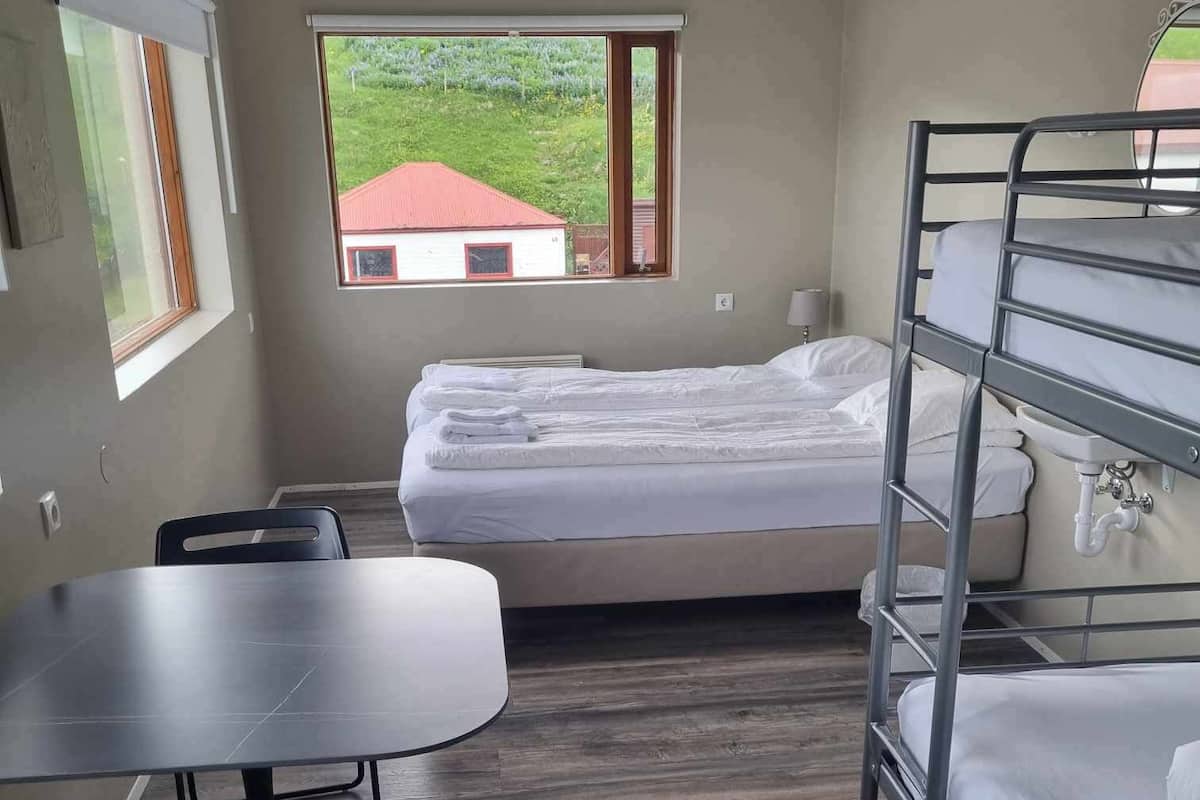 A compact room with a bunk bed, a single bed, a table, and chairs. There are windows providing a view of a red-roofed building and greenery outside.