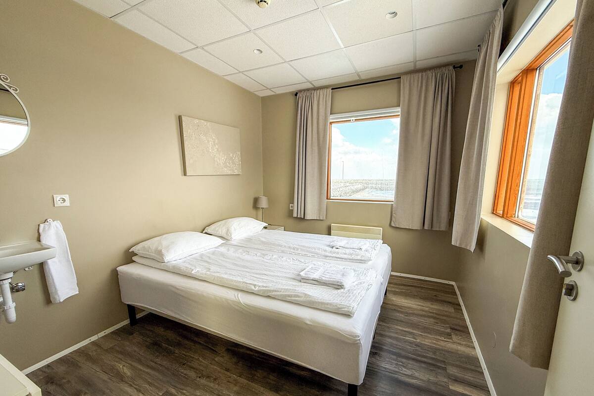 A hotel room with a bed, a bedside table, a lamp, a window with curtains, and a wall-mounted artwork.