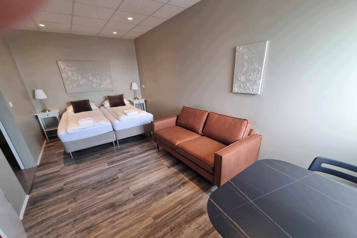 A modern hotel room with a wooden floor, a brown leather sofa, a bed with white linens, and a small table.