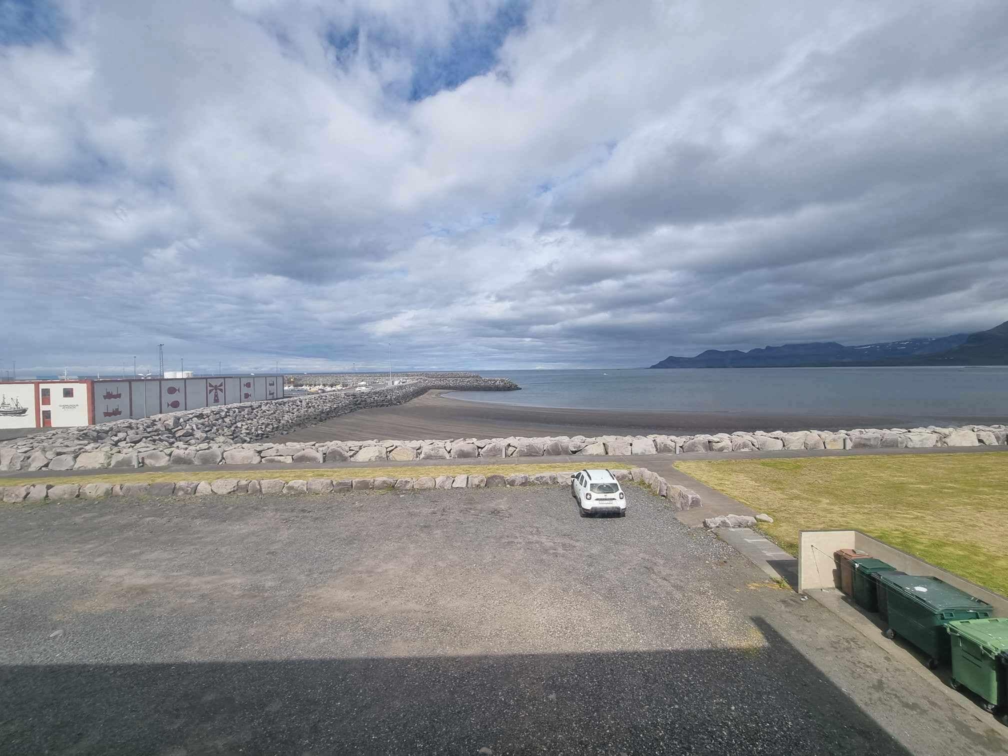 A parking area with a white car, a gravel path, and a view of a calm body of water with mountains in the distance.